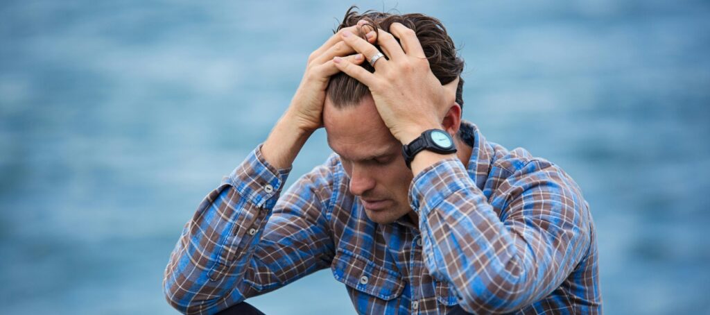 A man in a plaid shirt sits by the water looking distressed, symbolizing stress.